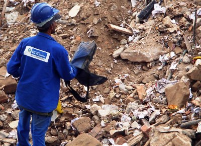 Homem com uniforme da Secretaria de Obras do Rio vasculha bolsa nos escombros que foram levados para entreposto